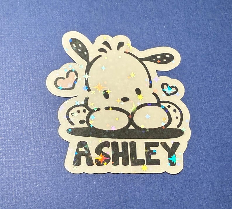 Puede incluir: Una pegatina hologr&aacute;fica con un perro de dibujos animados sobre un fondo brillante. El perro est&aacute; sentado con las patas cruzadas y tiene un coraz&oacute;n sobre su cabeza. La pegatina dice "ASHLEY" en letras negras.