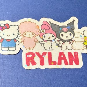 May include: A colorful sticker with five cartoon characters, including Hello Kitty, My Melody, Little Twin Stars, and Kuromi. The sticker also includes the name "RYLAN" in red letters.