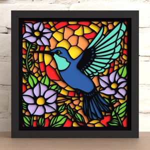 May include: A framed, stained-glass style artwork featuring a blue hummingbird in flight. The bird is surrounded by purple flowers, green leaves, and a colorful background of red, yellow, and orange. The artwork is in a black frame.