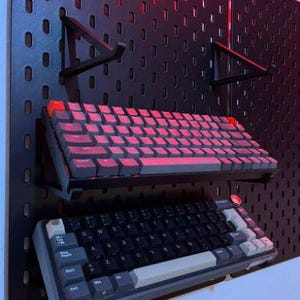 May include: Two keyboards mounted on a black pegboard wall. The top keyboard is a 60% keyboard with red keycaps. The bottom keyboard is a full-sized keyboard with black keycaps and a white space bar.