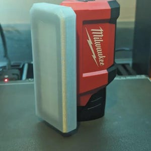 May include: A red and black Milwaukee power tool with a white rectangular attachment. The Milwaukee logo is visible in white. The items are on a dark gray surface.