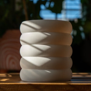 Puede incluir: Maceta de cerámica blanca con un diseño acanalado. La maceta tiene cinco canales y está sentada sobre una superficie de madera.