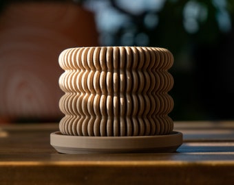 May include: A light brown, textured, cylindrical planter with a matching saucer. The planter has a wavy, rippled design.