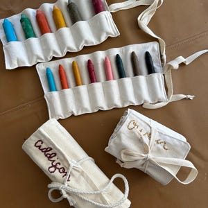 May include: Cream-colored crayon rolls with colorful crayons. Two rolls are open, displaying crayons in various colors. Two rolls are closed and tied with string, one embroidered with "Addison" and the other with a name.