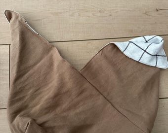 May include: A tan fabric bag with a pointed shape. The interior lining is white with a black plaid pattern. The bag is folded over, revealing the contrasting lining.