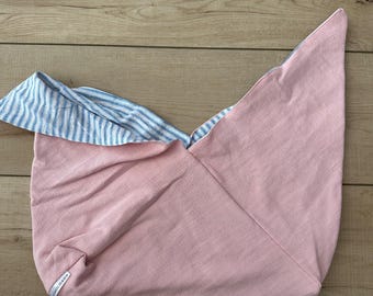 May include: A pink and blue striped baby carrier, made of soft fabric. The carrier is folded and appears to be unused, with a tag on the side. The carrier is designed to hold a baby.