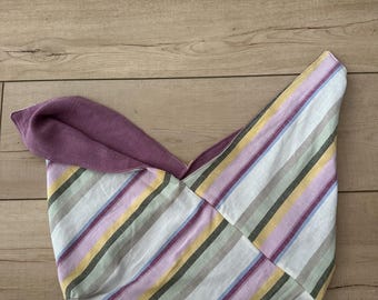 May include: A handmade tote bag with a striped pattern in pastel colors, including purple, yellow, green, and blue. The bag has a solid purple interior and is made of fabric. The bag is laying on a wooden surface.