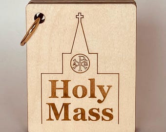 以下が含まれることがあります： セリフ体のフォントで「Holy Mass」という文字が書かれた明るい色の木製プラーク。プラークには、上部に十字架と円形のエンブレムが付いた教会の輪郭が描かれています。 金色のリングが左上隅に取り付けられています。