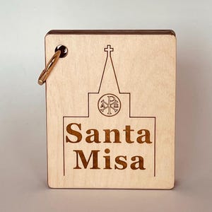 May include: A light-colored wooden keychain with a church illustration and the words "Santa Misa" engraved. The design includes a cross and a symbol within a circle. A gold-colored clasp is attached to the top left corner.
