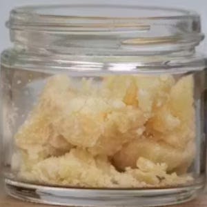 May include: A small, clear glass jar containing a light yellow, granular material. The jar has a wide opening and a clear lid. The substance inside appears to be solid, possibly a concentrate or extract.