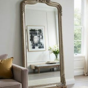 以下が含まれることがあります： 装飾的で銀色のフレームが付いた、大きく装飾的な全身鏡。鏡は白い壁に立てかけられており、ソファ、アートワーク、窓のある部屋を映しています。フレームには詳細な彫刻が施されています。