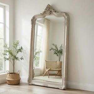 以下が含まれることがあります： オフホワイトのアンティーク調フレームの大きな全身鏡。白い壁に立てかけられています。フレームは上部と側面に詳細な彫刻が施されています。鏡には椅子と植物が映っています。鏡の高さは約180cmです。