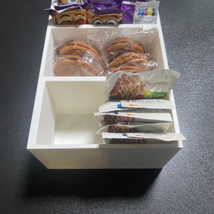 Puede incluir: Un organizador blanco de tres compartimentos con varios bocadillos. El compartimento superior contiene bolsas de aperitivos moradas y azules con ojos de dibujos animados. El compartimento del medio contiene varias galletas marrones en bolsas de plástico transparentes. El compartimento inferior contiene varias bolsas pequeñas de aperitivos con fondo blanco y texto azul y naranja.