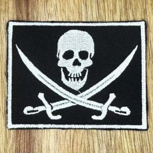 May include: Black rectangular patch featuring a white skull and crossbones design. Two white crossed swords are beneath the skull. The patch has a white border, set against a wooden background.