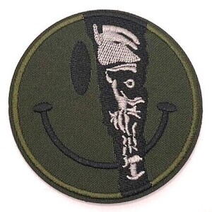 May include: A circular embroidered patch featuring an olive green smiley face design. The right side of the face is partially obscured by a vertical black stripe containing a white embroidered graphic of a figure wearing a hat and a coat.