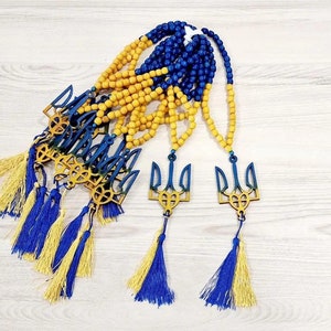 Puede incluir: Un conjunto de cuentas de oración de madera amarilla y azul con un dije de tridente. Las cuentas están enhebradas en un cordón y tienen un borla al final. El dije de tridente es un símbolo de Ucrania.