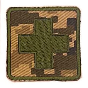 May include: A green medical cross patch on a camouflage background. The patch is square shaped with a green border.