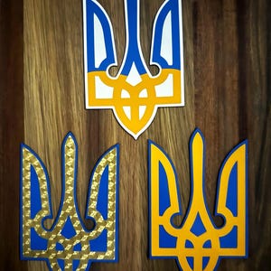 Könnte beinhalten: Drei ukrainische Tryzub-Embleme in Blau, Gelb und Weiß auf einer Holzoberfläche. Das obere Emblem ist ein durchgehendes Design, während die beiden unteren goldfarbene Hintergründe mit Mustern haben. Die Embleme sind ein Symbol der ukrainischen Nationalidentität.