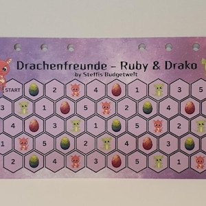 Könnte beinhalten: Ein lila Spielbrett mit dem Text "Drachenfreunde - Ruby & Drako" und "by Steffis Budgetwelt". Das Brett zeigt ein sechseckiges Raster mit Zahlen, Eiern und Zeichentrickdrachen. Das Spielbrett hat oben eine Reihe von Löchern.