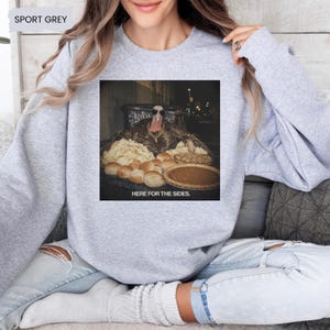 May include: A sport grey crewneck sweatshirt featuring a graphic of a turkey wearing sunglasses and a gold chain, sitting behind a table filled with dinner rolls and a pie, with the text HERE FOR THE SIDES printed below the image.