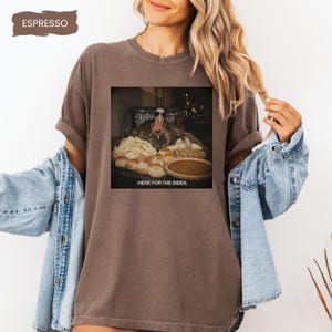 May include: An espresso-colored t-shirt featuring a graphic of a turkey wearing sunglasses and a gold chain, surrounded by various side dishes. The text below the turkey reads, "HERE FOR THE SIDES." The shirt has a crew neckline and short sleeves.