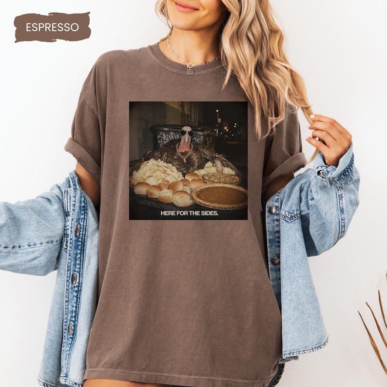 May include: An espresso-colored t-shirt featuring a graphic of a turkey wearing sunglasses and a gold chain, surrounded by various side dishes. The text below the turkey reads, "HERE FOR THE SIDES." The shirt has a crew neckline and short sleeves.