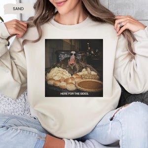 May include: A sand-colored crewneck sweatshirt featuring a graphic print of a turkey wearing sunglasses and a gold chain, surrounded by Thanksgiving side dishes including mashed potatoes, rolls, and a pie, with the text HERE FOR THE SIDES.