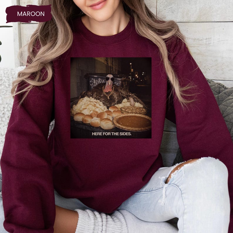 May include: A maroon crewneck sweatshirt featuring a graphic print of a turkey wearing sunglasses and a gold chain, surrounded by various side dishes including rolls and a pie, with the text "HERE FOR THE SIDES." printed below the image.