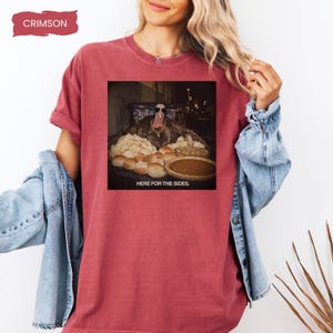 May include: A crimson short-sleeve t-shirt featuring a graphic print of a turkey wearing sunglasses behind a table of food with the text "HERE FOR THE SIDES." The shirt has a crew neckline and a relaxed fit, with a small "CRIMSON" label in the corner.