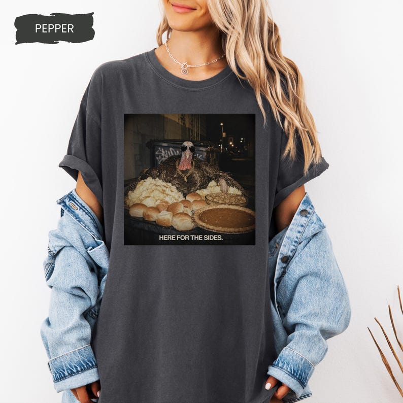 May include: A charcoal gray short-sleeve t-shirt featuring a graphic print of a turkey wearing sunglasses behind a table filled with various side dishes and a pie, with the text "HERE FOR THE SIDES." printed in white capital letters below the image.