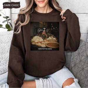May include: A dark chocolate brown crewneck sweatshirt featuring a graphic print of a turkey wearing sunglasses behind a table filled with various side dishes and a pie. The text below the image reads, "HERE FOR THE SIDES."