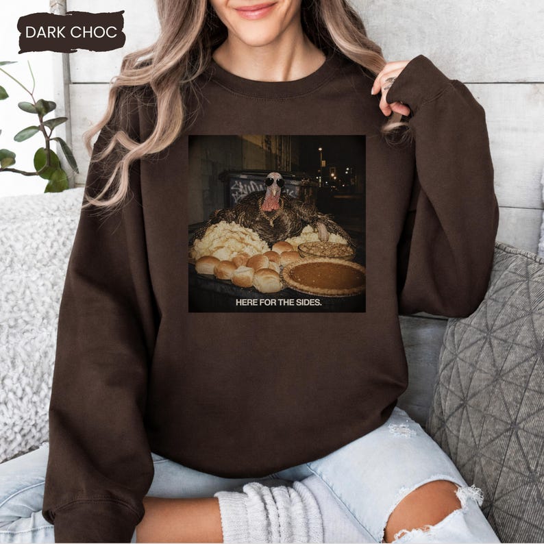 May include: A dark chocolate brown crewneck sweatshirt featuring a graphic print of a turkey wearing sunglasses behind a table filled with various side dishes and a pie. The text below the image reads, "HERE FOR THE SIDES."