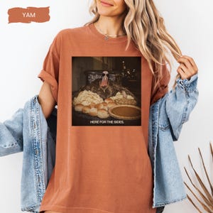 May include: A rust-colored short-sleeve t-shirt featuring a graphic print of a turkey wearing sunglasses and a gold chain, surrounded by various side dishes. The text "HERE FOR THE SIDES." is printed below the image. The shirt has a crew neckline.