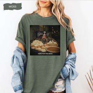 May include: A moss green short-sleeve t-shirt featuring a graphic print of a turkey wearing sunglasses behind a table of food with the text HERE FOR THE SIDES. The shirt has a crew neckline and a relaxed fit, showcasing a casual style.