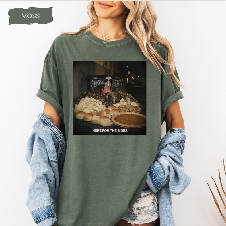 May include: A moss green short-sleeve t-shirt featuring a graphic print of a turkey wearing sunglasses behind a table of food with the text HERE FOR THE SIDES. The shirt has a crew neckline and a relaxed fit, showcasing a casual style.