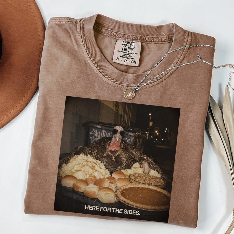 May include: A tan short-sleeve t-shirt featuring a graphic print of a turkey wearing sunglasses and a gold chain, surrounded by mashed potatoes, rolls, and a pie. The text below the image reads, "HERE FOR THE SIDES." The shirt has a Comfort Colors label.