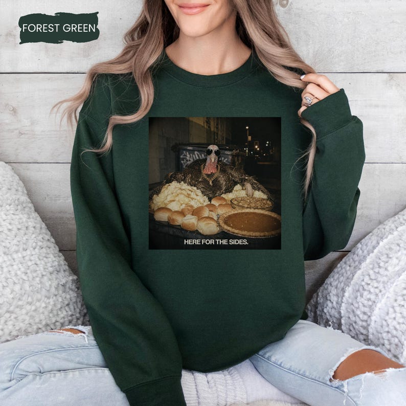 May include: A forest green crewneck sweatshirt featuring a graphic print of a turkey wearing sunglasses and a gold chain, surrounded by various side dishes including rolls and a pie, with the text "HERE FOR THE SIDES." printed below the image.