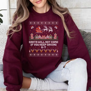May include: Burgundy sweatshirt with a festive Christmas design. The design includes Santa in his sleigh, reindeer, houses, and the text "SANTA WILL NOT COME IF YOU KEEP SAYING 6-7". The design is in red, white, and gold.