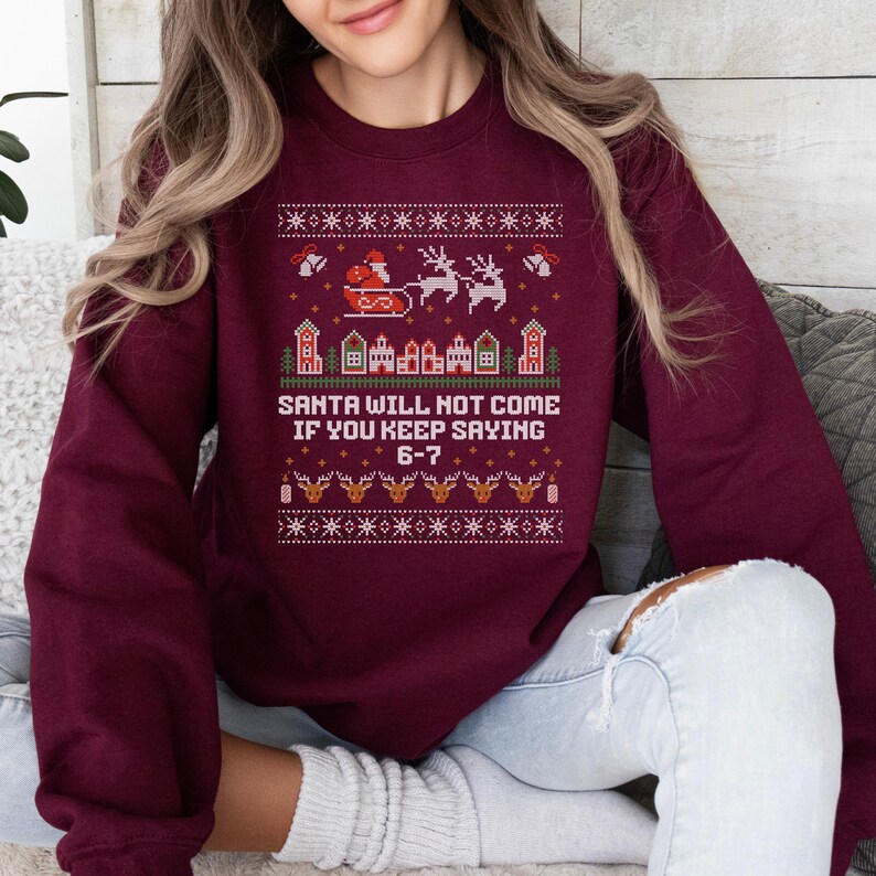 May include: Burgundy sweatshirt with a festive Christmas design. The design includes Santa in his sleigh, reindeer, houses, and the text "SANTA WILL NOT COME IF YOU KEEP SAYING 6-7". The design is in red, white, and gold.