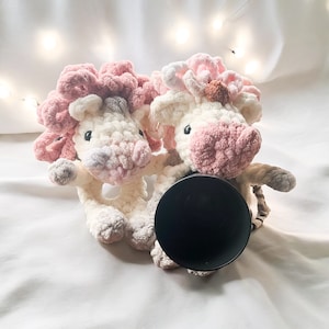 May include: Two pink and white crocheted unicorn plushies with a black camera lens in the centre. The plushies have pink and white yarn and are holding the lens.