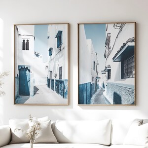 May include: Two framed prints depict a narrow, sunlit alleyway with white buildings and blue accents. The artwork features a clear blue sky and a sense of depth, creating a serene and inviting atmosphere. The frames are a warm gold color.