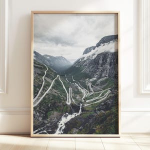 May include: A framed print featuring a winding mountain road through a valley. The road snakes through the green landscape, with waterfalls and rocky cliffs. The sky is overcast with clouds, and the frame is a light wood color.
