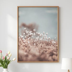 Op de afbeelding: Een ingelijste kunstdruk met een veld witte, pluizige bloemen tegen een zachte roze en blauwe achtergrond. Het kunstwerk is gepresenteerd in een goudkleurige lijst, wat een serene, natuurlijke esthetiek biedt voor interieurdecoratie.