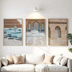 May include: Three framed prints featuring architectural scenes. One shows a tiered fountain with turquoise water, another a white fountain in a courtyard, and the third a building facade with ornate windows. The prints are displayed above a white sofa.