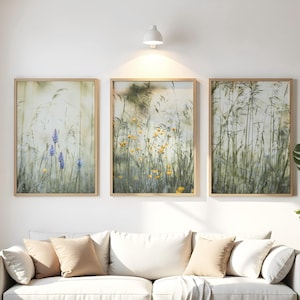 Może przedstawiać: Trzy oprawione wydruki artystyczne przedstawiające zbliżenia polnych kwiatów i wysokich traw na polu. Wydruki mają naturalną, neutralną paletę kolorów z miękkimi zieleni, żółciami i fioletami. Ramki są w jasnym kolorze drewna.