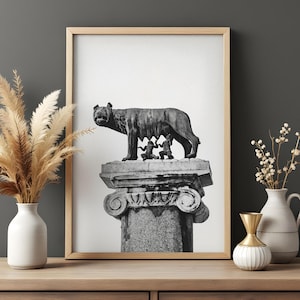 May include: Black and white framed print of the Capitoline Wolf statue. The sculpture depicts a she-wolf nursing Romulus and Remus, the legendary founders of Rome. The artwork is displayed on a wooden surface with decorative vases.