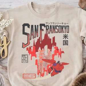 May include: Beige sweatshirt featuring a red and black graphic of a city skyline and a flying robot. The text "SAN FRANSOKYO" is displayed above the skyline, with Japanese characters and the words "BIG HERO" below.