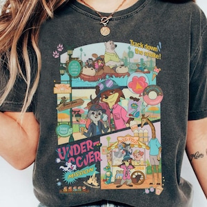 May include: Dark heather gray t-shirt featuring a colorful cartoon graphic with the text "Undercover Mission" and "Track down the snake!". The graphic includes various animal characters and food items. The shirt is a comfortable, casual style.