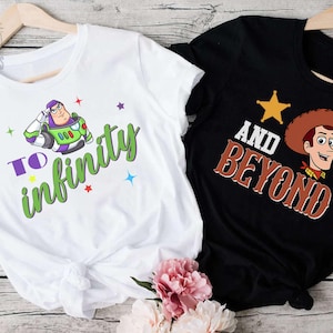 May include: Two t-shirts, one white and one black, with cartoon graphics. The white shirt features Buzz Lightyear with the text "To Infinity" in green and purple. The black shirt displays Woody with the text "And Beyond" in brown.