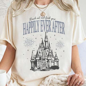 以下が含まれることがあります： 「Reach out and find your HAPPILY EVER AFTER」の文字と、お城の白黒イラストがプリントされたクリーム色のTシャツ。 ゆったりとしたフィット感で半袖です。白いマグカップが左手に持たれています。